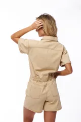 Mono corto color beige, estilo denim, con cuello camisero, cierre frontal con botones, mangas cortas y bolsillos en el pecho y laterales.
