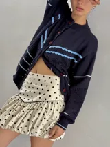 Saco de punto calado color beige claro, con cuello redondo y cierre frontal de botones azules. Presenta detalles decorativos en crochet verde y negro en el frente y puños.