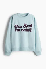 Buzo de tejido de punto color celeste claro, con cuello redondo y manga larga. Presenta un estampado frontal con el texto "New York EST. 1985 5TH AVENUE" en tipografía universitaria color bordó.