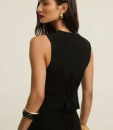 Chaleco negro de sastrería con cuello redondo, cierre frontal con botones metálicos y lazo cruzado en la espalda.