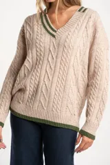Sweater de lana y nylon color beige jaspeado, con diseño de punto trenzado (cable knit) en todo el cuerpo y mangas. Presenta cuello en V con ribete de dos franjas verdes y puños y bajo con ribete verde liso.
