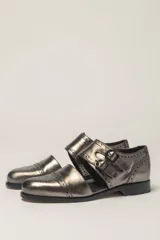 Zapato bajo de cuero negro, estilo monk strap con dos hebillas ajustables sobre el empeine. Presenta detalles de brogue (perforaciones) en la puntera y alrededor del borde superior. Tiene un taco bajo y ancho.