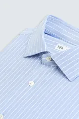 Camisa celeste a rayas verticales blancas, de corte recto, con cuello italiano abierto, mangas largas con puños abotonados y cierre frontal con botones.