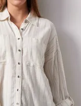 Camisa de lino a rayas verticales color crema, con cuello con solapa, abertura central con botones, bolsillos delanteros y mangas 3/4 regulables con presilla y botón.
