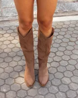 Botas texanas de caña media confeccionadas en cuero gamuzado color marrón. Presentan un diseño de corte clásico con costuras decorativas en el empeine, puntera levemente redondeada y taco bajo de madera.