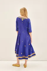 Vestido largo azul vibrante con escote en V profundo, mangas 3/4 y caída fluida. Presenta un elaborado bordado frontal en el escote con piedras, cintas e hilos de lurex en tonos dorados y plateados, con pequeños apliques florales dorados distribuidos en el cuerpo de la prenda. Incluye un accesorio colgante de cuentas doradas a modo de bufanda o detalle decorativo.