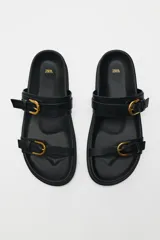 Sandalias planas negras con dos tiras anchas y hebillas doradas.