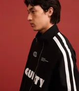 Campera deportiva negra con cuello alto y cierre frontal. Presenta franjas blancas contrastantes a lo largo de las mangas y detalles gráficos en blanco en el pecho, incluyendo la palabra "GUILTY" en grande.