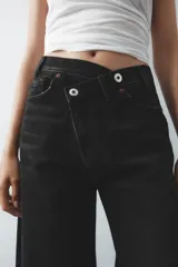 Pantalón de jean celeste de tiro alto, corte wide leg y largo hasta el tobillo. Presenta un diseño asimétrico en la cintura con cierre frontal cruzado con cremallera y botones metálicos.