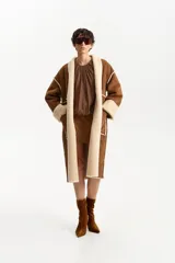 Abrigo largo de diseño envolvente, exterior de cuero gamuzado color marrón y forrería interior de corderito color beige. Presenta solapas anchas de corderito y puños también de corderito.