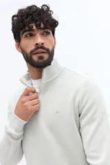 Buzo de algodón color verde musgo, con cuello alto y cierre frontal parcial. Presenta puños y cintura acanalados, y un pequeño logo bordado en el pecho.