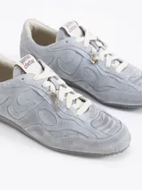 Zapatilla de cuero color marrón oscuro con efecto gastado y apliques de gamuza con diseño de llamas o remolinos. Posee cordones color bordó y un dije metálico en forma de llave colgando de los cordones.