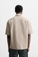 Sobrecamisa color beige de tejido elástico, con cuello solapa, manga corta y bolsillos de parche en el pecho. Cierre frontal con botones a presión.