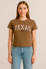 Remera marrón de algodón con cuello redondo y mangas cortas. Estampado frontal con la palabra "Texas" en color rosa con borde negro y el año "1985" debajo.