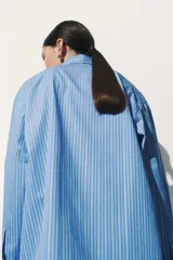 Camisa celeste y blanca a rayas verticales, de corte amplio y mangas largas con puños abotonados. Cuello clásico y cierre frontal con botones.