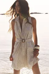 Vestido corto color beige con bordados florales perforados, cuello solapa, sin mangas, cinturón de tela con hebilla redonda y cierre frontal con botones.