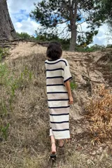 Vestido tejido artesanalmente en hilado 70% algodón y 30% acrílico, con cuello redondo y mangas cortas. Diseño a rayas horizontales en tonos crudo, naranja y beige. Corte recto con aberturas laterales en el ruedo.