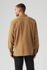 Camisa color marrón claro de corte clásico, con cuello abotonado, cierre frontal con botones, dos bolsillos de parche con botón en el pecho y mangas largas con puños abotonados.