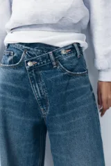 Pantalón de jean celeste de tiro alto, corte wide leg y largo hasta el tobillo. Presenta un diseño asimétrico en la cintura con cierre frontal cruzado con cremallera y botones metálicos.