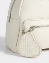 Mochila pequeña de cuerina color rosa pálido, con moño en relieve en el frente y llavero con forma de corazón trenzado. Tiene correas regulables, manija superior, cierre superior, bolsillo frontal con cierre y bolsillos laterales.