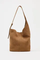 Bolso tipo saca asimétrica de serraje color marrón. Tiene asa bandolera ajustable y cierre con imán. Incluye cartera con bolsillo extraíble.