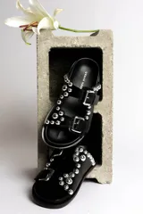 Sandalia tipo mule negra de cuero, con dos tiras ajustables con hebillas metálicas plateadas y apliques de tachas esféricas plateadas. Presenta un borde festoneado en la tira superior y suela de plataforma negra.