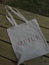 Bolso tipo tote de tela color crema con asas largas y logo de la marca Meilé estampado en color rosa.