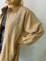 Camisola larga color beige con apliques de strass plateados, cuello camisero, mangas largas con puños abotonados y cierre frontal con botones. Puede usarse como vestido o sobrecamisa.