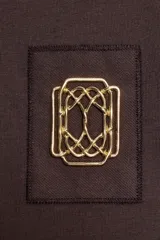 Camiseta marrón de algodón de manga corta y cuello redondo. Presenta un bolsillo en el pecho con un broche metálico dorado con diseño intrincado.
