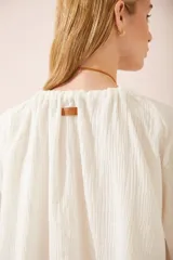 Camisa oversize blanca de lino, con cuello en V, mangas largas abullonadas con puños elásticos y lazos decorativos.