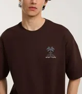 Remera marrón de corte relajado, confeccionada en algodón. Presenta cuello redondo, mangas cortas y estampa frontal y trasera de palmeras con la leyenda 'Work Hard'.