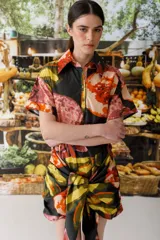 Vestido camisero corto con estampado abstracto en tonos naranja, rosa, amarillo y negro. Tiene cuello camisero, mangas cortas y cierre frontal con botones. Incluye lazo en la cintura para ajustar.