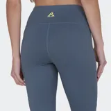 Calza deportiva de tiro alto, color gris azulado, con recortes laterales en amarillo claro. Presenta paneles con microperforaciones en la zona de la cadera y una franja vertical amarilla en la parte inferior de las piernas.