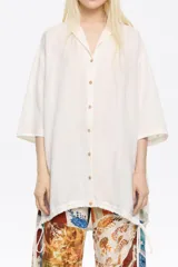 Camisa larga color blanco, de corte oversize y escote con solapas. Tiene manga corta recta y ancha, cierre con botonadura frontal y bajo ajustable con cordones de tejido.