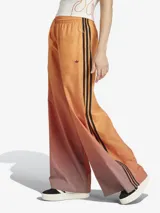 Pantalón deportivo Adidas de corte holgado con diseño degradado en tonos naranja y marrón. Presenta las tres rayas negras características de la marca en los laterales y logo bordado en la parte superior.