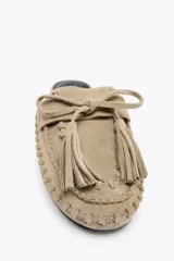 Zapato plano destalonado tipo mule, color beige, confeccionado en serraje. Presenta antifaz y lazo con borlas en la parte delantera.