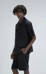 Camisa negra de manga corta estilo técnica, confeccionada en microfibra liviana. Tiene doble bolsillo frontal con cierre de cremallera y broches en lugar de botones.