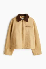 Campera color beige de algodón Oxford con cuello de pana marrón en contraste. Modelo de corte holgado con cierre frontal, hombros caídos, bolsillos laterales ribeteados y bolsillo superior con cierre. Ajuste con botones a presión en los puños y en la parte posterior del bajo.