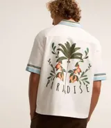 Camisa masculina de algodón con corte boxy, cuello con solapa y rayas, abotonado delantero, bolsillo con estampa floral y estampa trasera de palmera con la palabra 'Paradise'.
