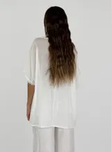 Blusa gris de lino y algodón, de corte amplio, con cuello redondo y mangas cortas con terminación desflecada. Presenta un volante diagonal en el frente.