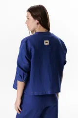 Chaqueta color crudo de lino con cuello redondo, manga tres cuartos, cierre frontal con botones y bolsillos de parche. Detalle de costuras en contraste color marrón.