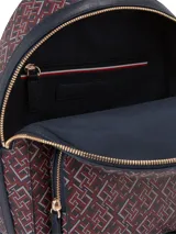 Mochila de silueta clásica con estampado integral de monograma en tonos marrón oscuro y blanco. Presenta un compartimento principal con cierre, un bolsillo exterior con cremallera y un asa superior negra. El logo TH metálico se encuentra centrado en el frente.