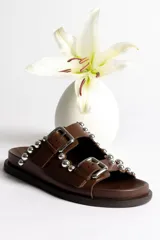 Sandalia tipo mule negra de cuero, con dos tiras ajustables con hebillas metálicas plateadas y apliques de tachas esféricas plateadas. Presenta un borde festoneado en la tira superior y suela de plataforma negra.