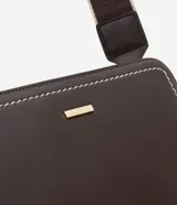 Cartera transversal de material sintético color marrón oscuro, de forma rectangular y estructura rígida. Presenta pespuntes decorativos en color blanco en los bordes frontales y cierres metálicos dorados. Incluye un asa ancha desmontable con diseño de rayas beige y marrón.