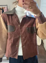 Chaqueta marrón con mangas de piel sintética color beige, cuello y solapa de cuero sintético marrón oscuro, cierre con botones dorados y bolsillos frontales con solapa de cuero sintético marrón oscuro.