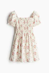 Vestido corto de popelina de algodón color blanco con estampado floral en tonos rosados. Tiene escote cuadrado con elástico, mangas cortas abullonadas con elástico y cuerpo fruncido.