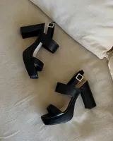 Sandalias negras de cuero sintético con plataforma y tacón cuadrado. Tienen dos tiras anchas sobre el empeine y una tira con hebilla alrededor del tobillo.