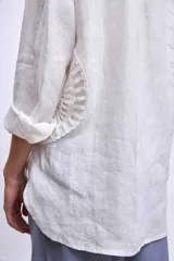 Camisa blanca de lino con cuello clásico, cierre frontal con botones y mangas largas con puños abotonados. Presenta apliques de encaje en hombros y bolsillos delanteros.