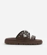 Sandalias planas marrones con punta redonda, tiras anchas con tachas plateadas y hebilla metálica estilo western.