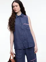 Camisa sin mangas negra de lino con cuello clásico, cierre frontal abotonado y bolsillo de parche en el pecho. Presenta bordados tipo pespunte en contraste en el frente y canesú trasero.